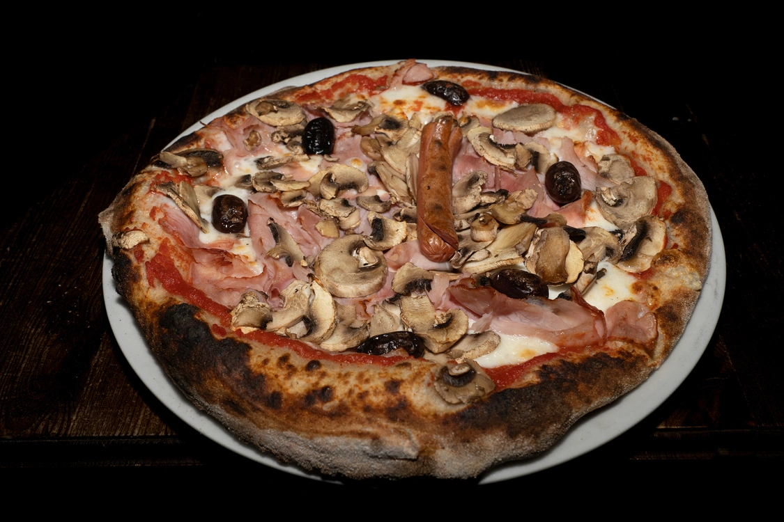 Pizza Capricciosa artigianale con base pomodoro e fior di latte, farcita con prosciutto cotto, funghi freschi affettati, olive nere e un wurstel centrale, cotta in forno a legna — Pizzeria Alla Botte, Misilmeri.